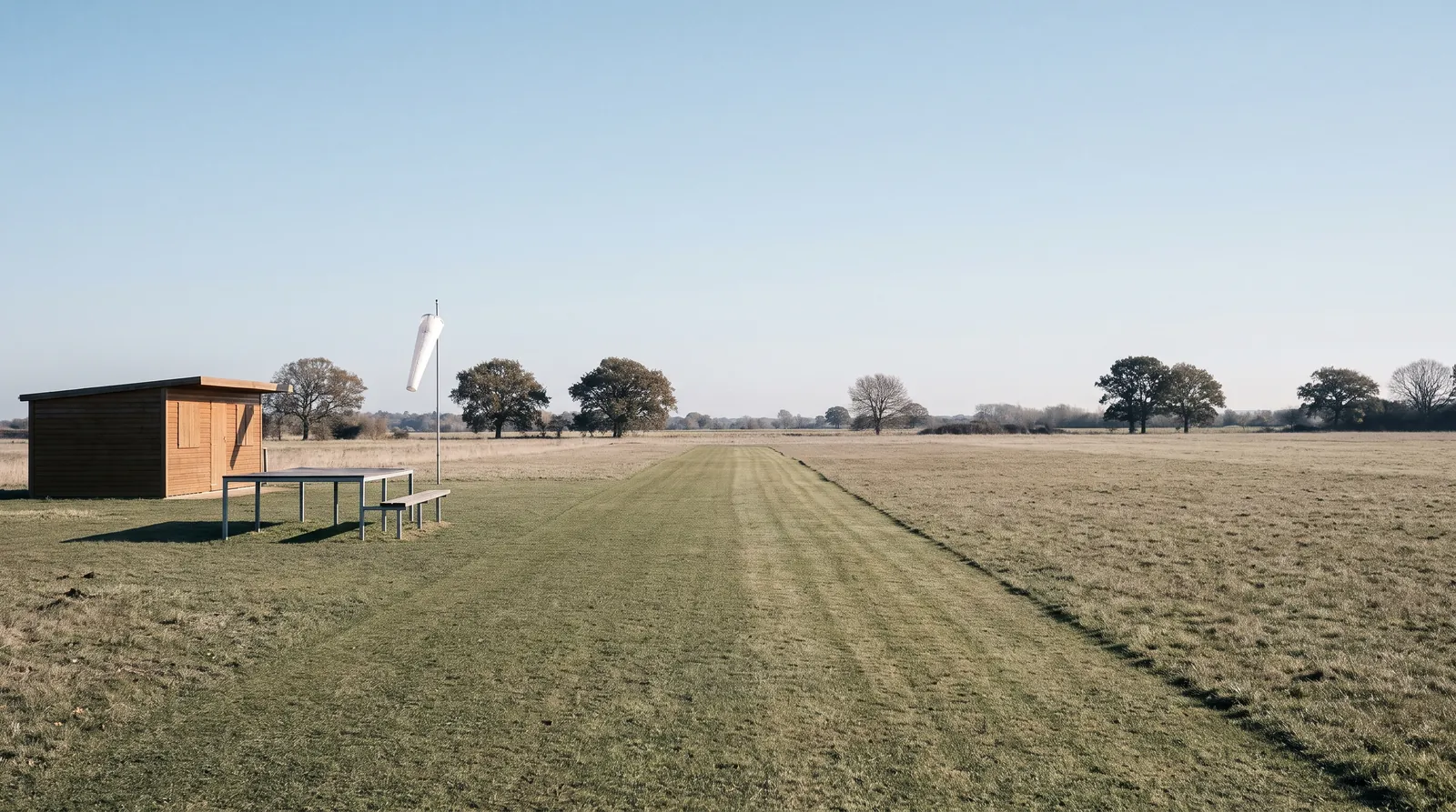 Campo de vuelo de aeromodelismo vacío con una pista de hierba segada, una caseta de madera y encinas al fondo.