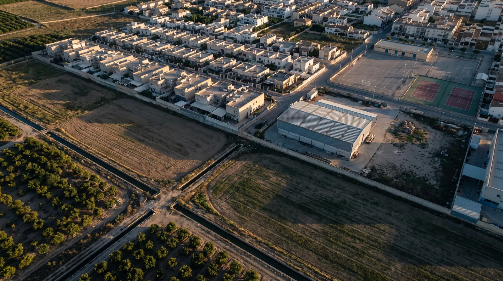 Huertas y campos abiertos en el borde de una localidad con casas bajas, una nave industrial y pistas deportivas vacías.